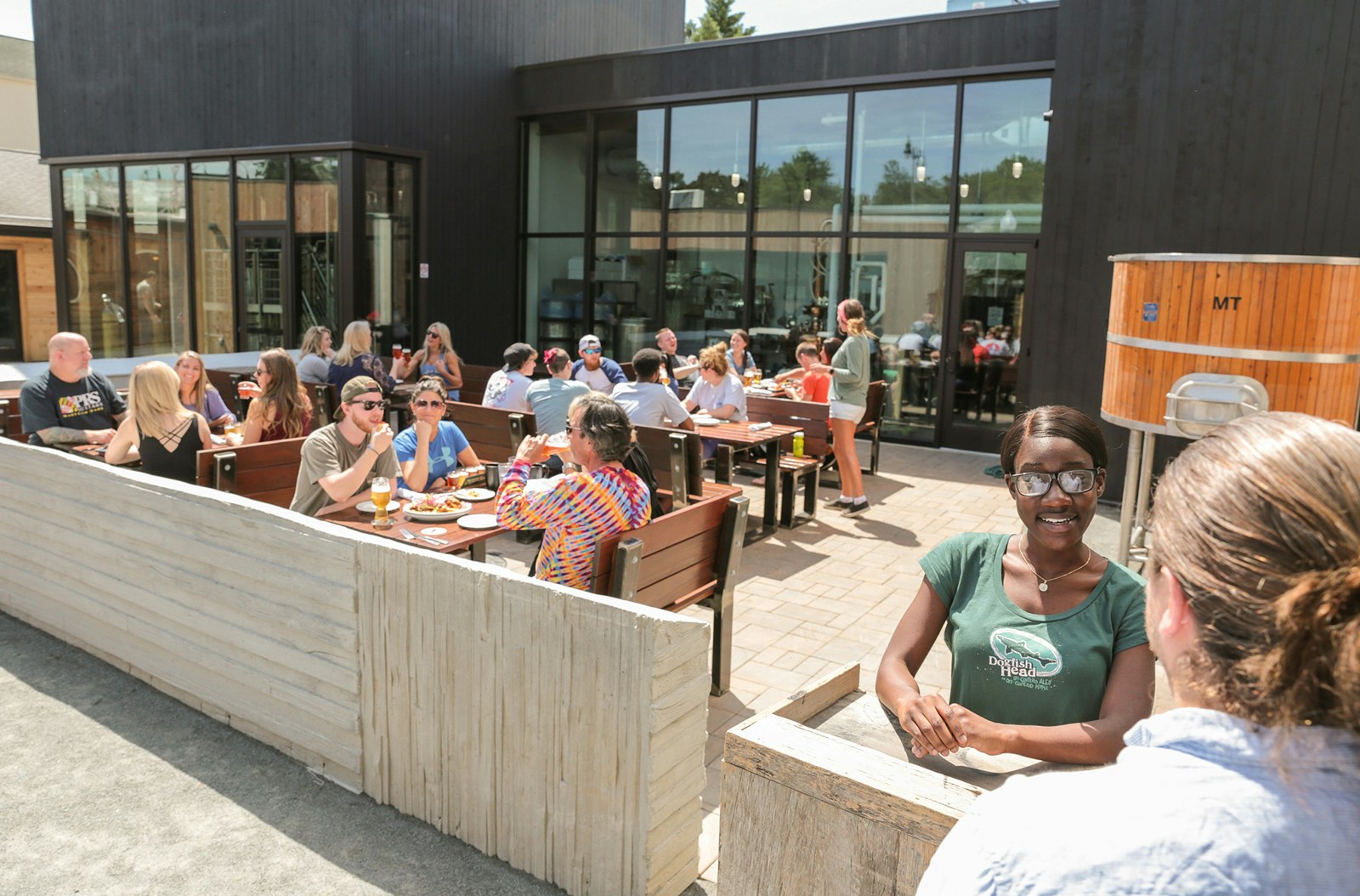 Hostess checks in a guest at Dogfish Brewing in Delaware, with a modern, concrete patio full of people in the background. Many craft breweries in the US now feature outdoor patio spaces. 