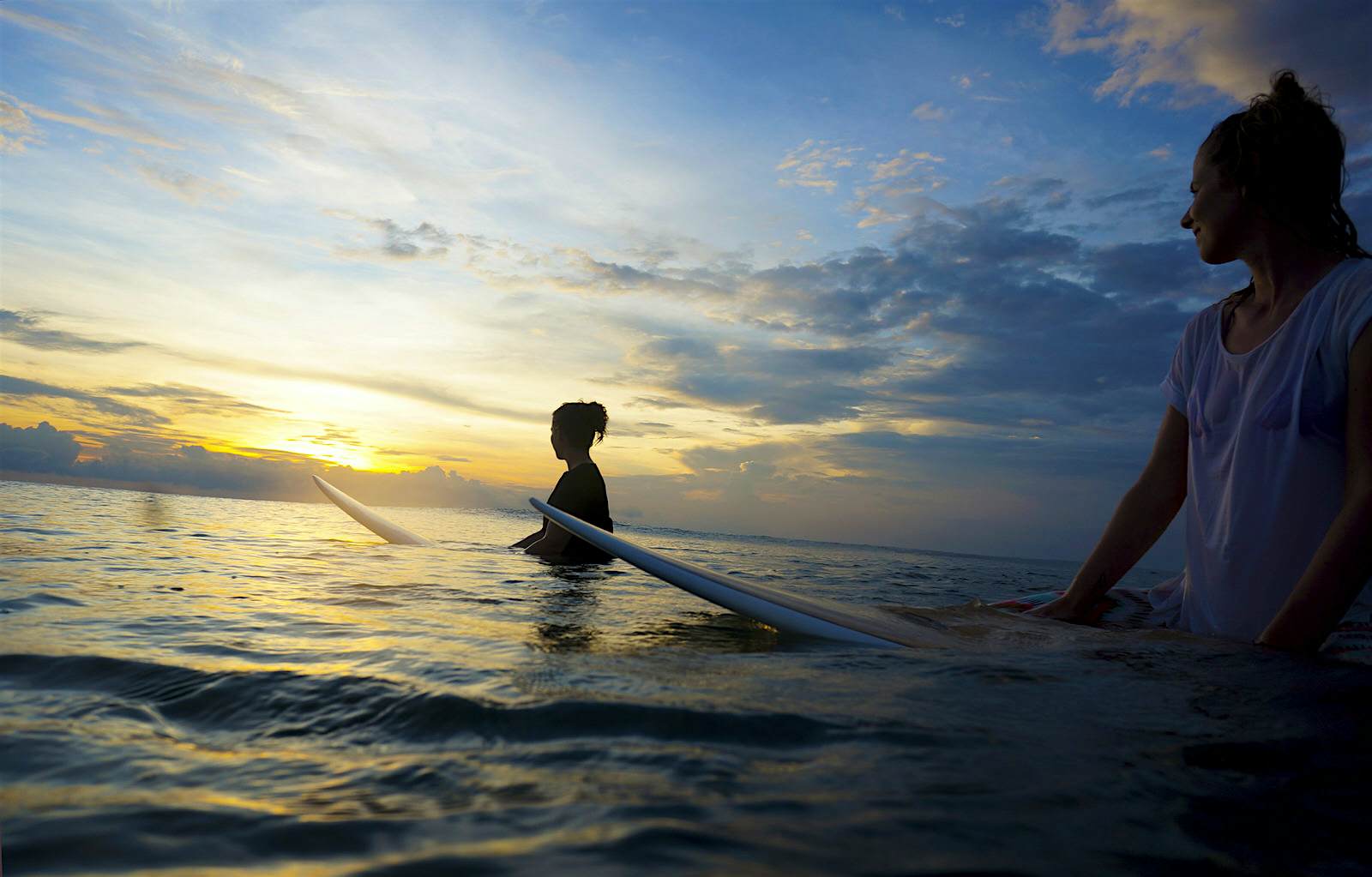 Features - Indonesia, Bali, Canggu, two female surfers in the water watching the sun Two female surfers sit on their boards in calm water looking towards the setting sun © Westend61 / Getty Images