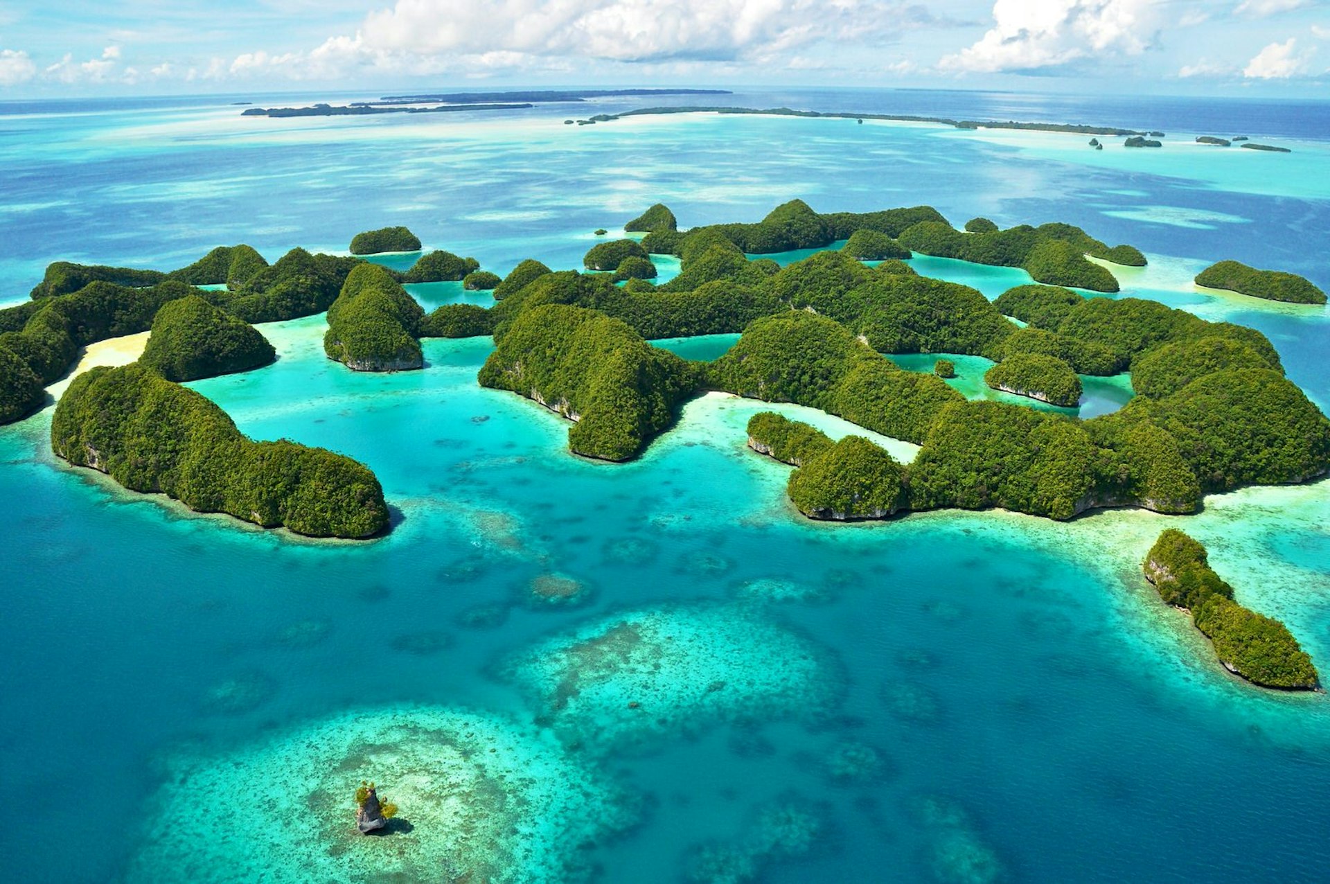 An aerial shot of the islands of Palau. The small lumpy islands are covered in dense greenery and surrounded by jewel-blue waters. 