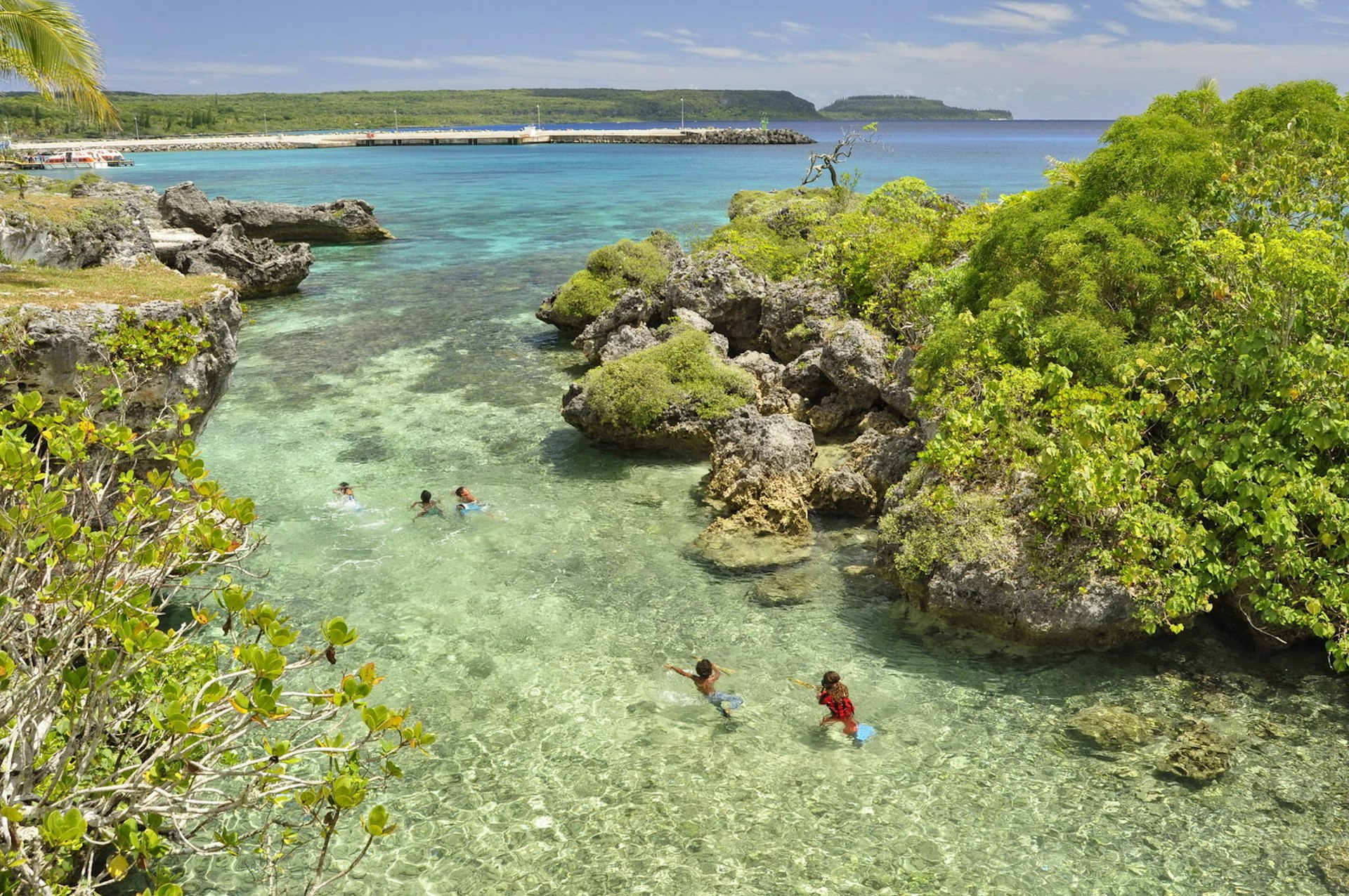 Lagoon on Mare Island, New Caledonia. There are several people paddling in the clear water. Either side of the lagoon are hulking grey rocks topped with vibrant green plants and moss. 
