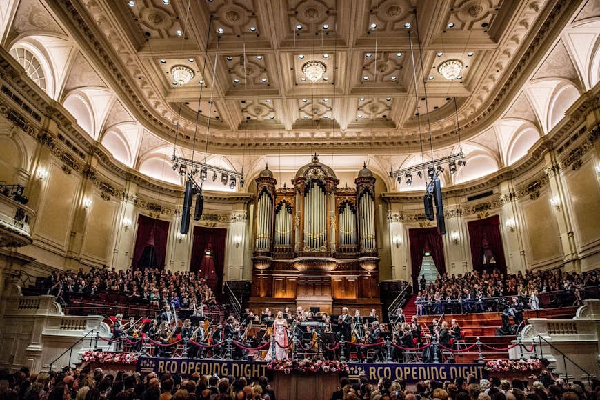 The grand interior of the Concertgebouw orchestra hall, with a full orchestra present on the stage, including a huge organ, and a large crowd filling the stalls