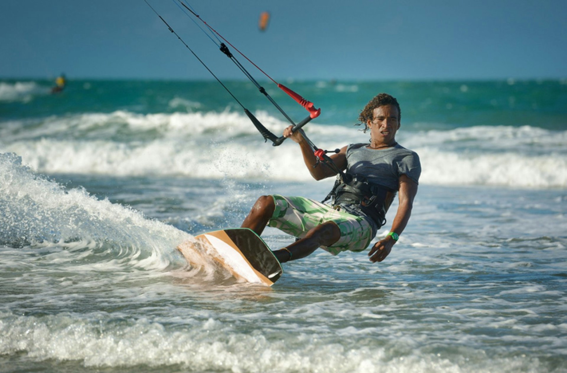 A male kitesurfer with long hair leans away from his board with one hand holding on to the handle of his kite equipment