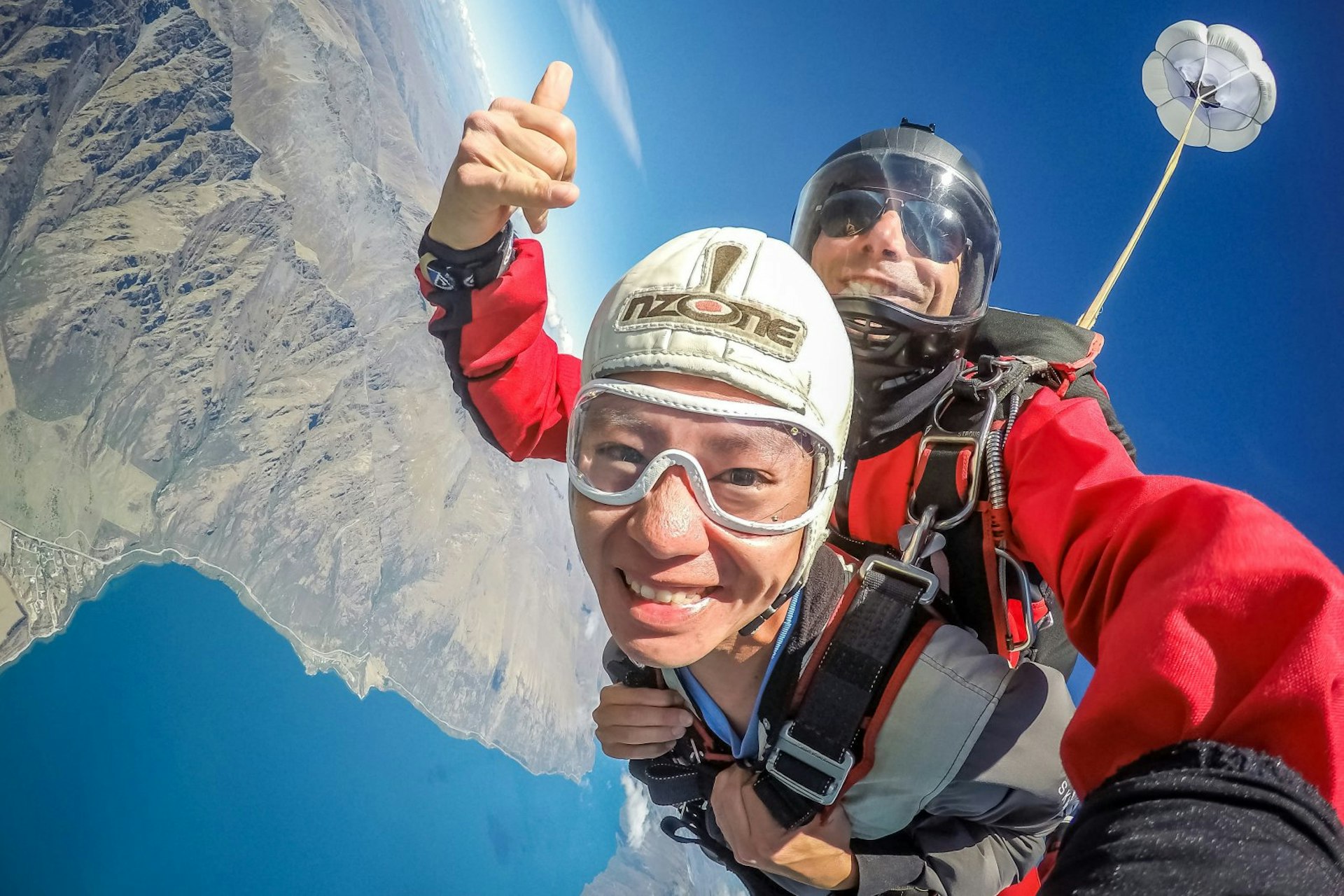 The skydiver smiles at the camera during freefall, while the instructor gives a thumbs up. The land and lake can be seen below