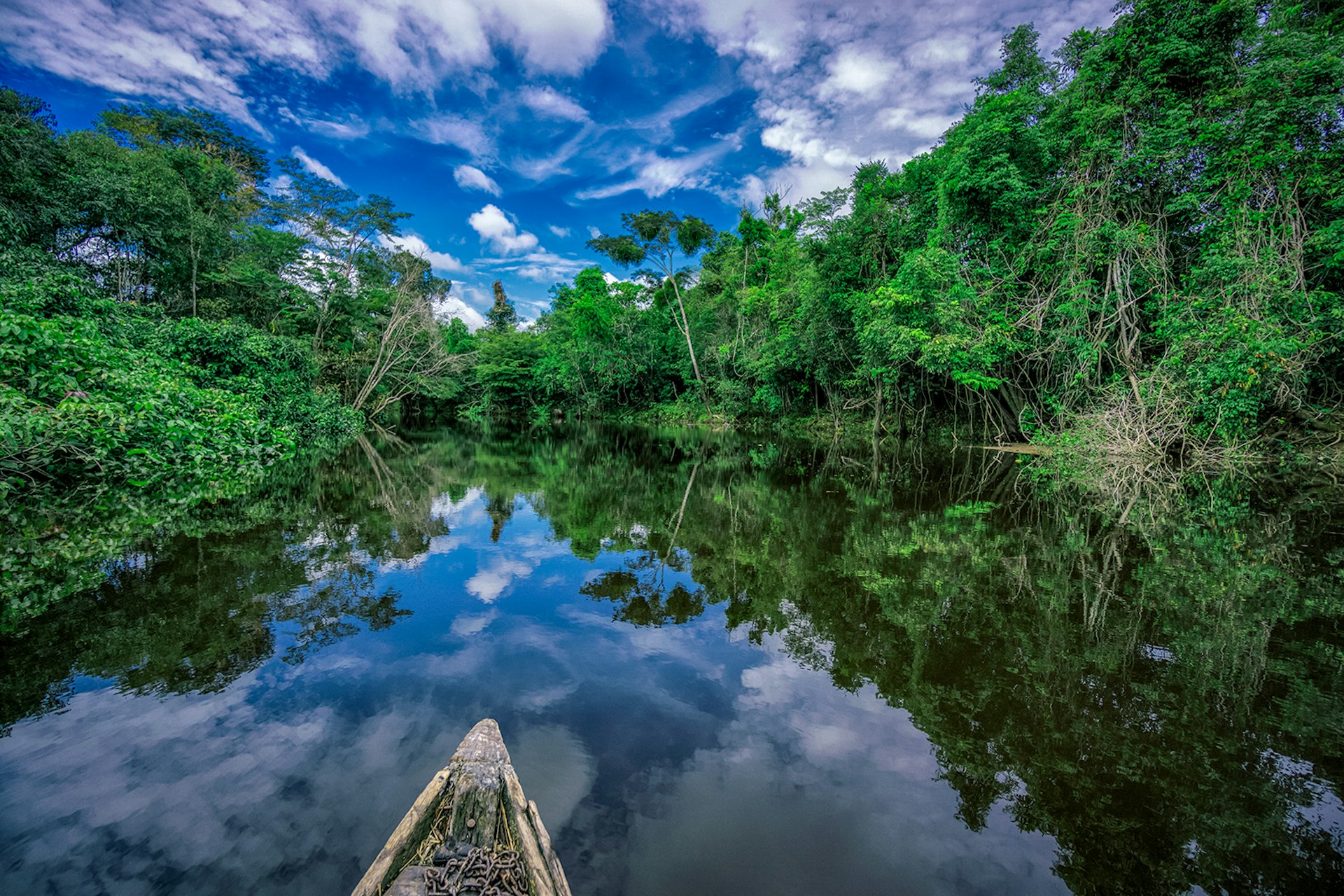 A wooden canoe drifts down a glassy Nanay river with Amazon forest on both shores. The photo is taken from the body of the canoe, with just the tip in frame. The cloudy blue sky is reflected in the still water.