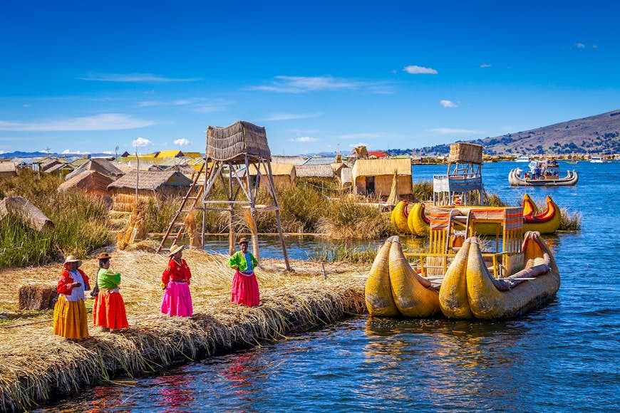 Four women in brightly colored, traditional skirts stand on a shoreline close to two curved boats made out of reeds