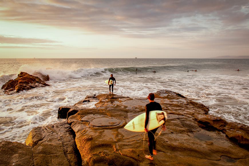 Two surfers walk out onto a rust-colored rock towards the surf at sunset. There are several other surfers already in the sea.
