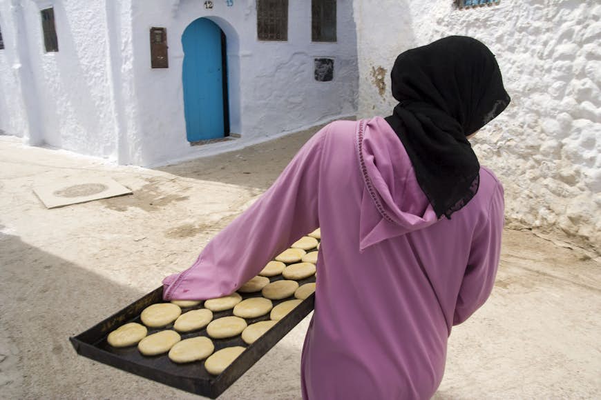 Young female baker dressed on a pink hijab carrying on a metallic tray some recently made cookies to a client, Chefchaouen, Morocco. Image by Santiago Urquijo / Getty Images