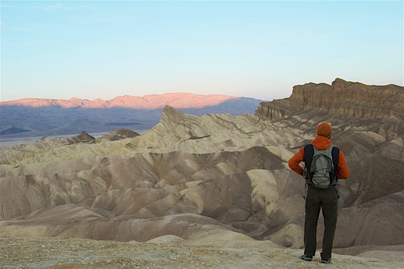 Features - GettyImages-172455557_medium Sunrise in Death Valley National Park. A person stands with their back to the camera, they are wearing a woolly orange hat, an orange jumper, a grey backpack and grey trousers. They look over the undulating sandstone structures of Death Valley, the pink morning light has just started to touch the furthest peak.