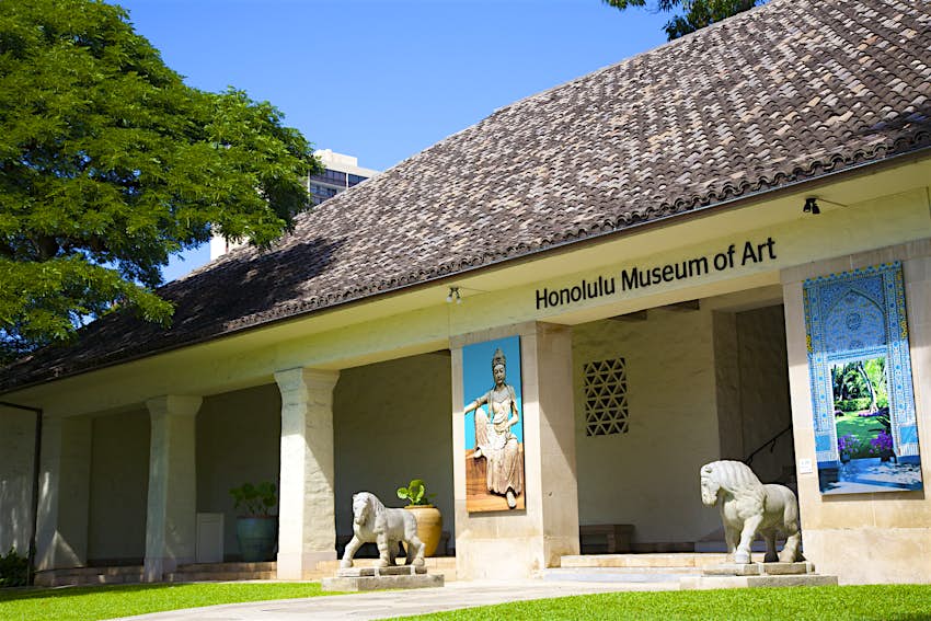 The outside of the Honolulu Museum of Art. The building is light colored with a series of small stone pillars and a dark brown terra-cotta roof. There are a pair of horse statues at the entrance and a big tree towards the end of the building; hidden Honolulu