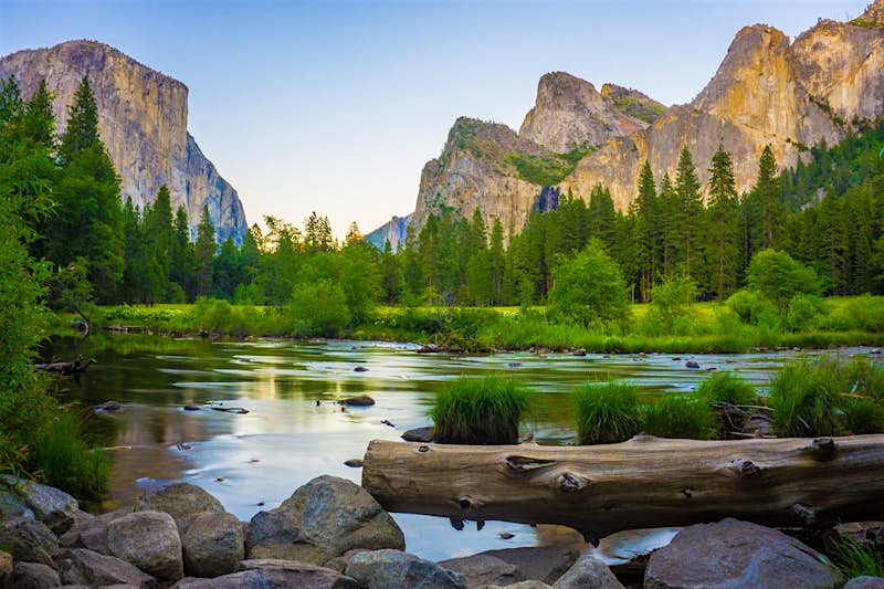 Features - GettyRF_535829001 Valley view of Yosemite National Park. Huge rugged mountains are set against a clear blue sky, with tall pine trees and a lake in the foreground.