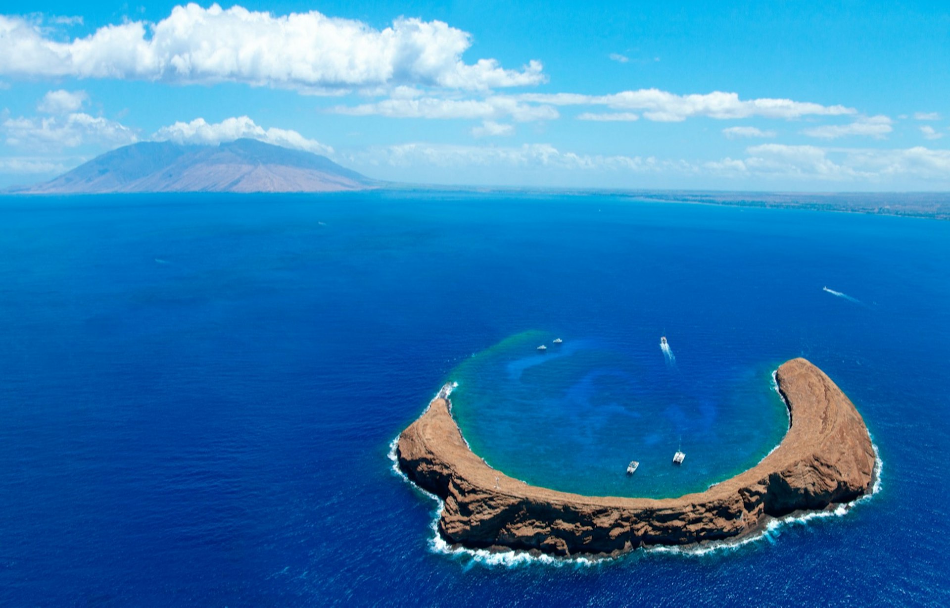 Aerial view of the Molokini Crater in a deep blue ocean. West Maui can be seen in background; Maui travel experiences 