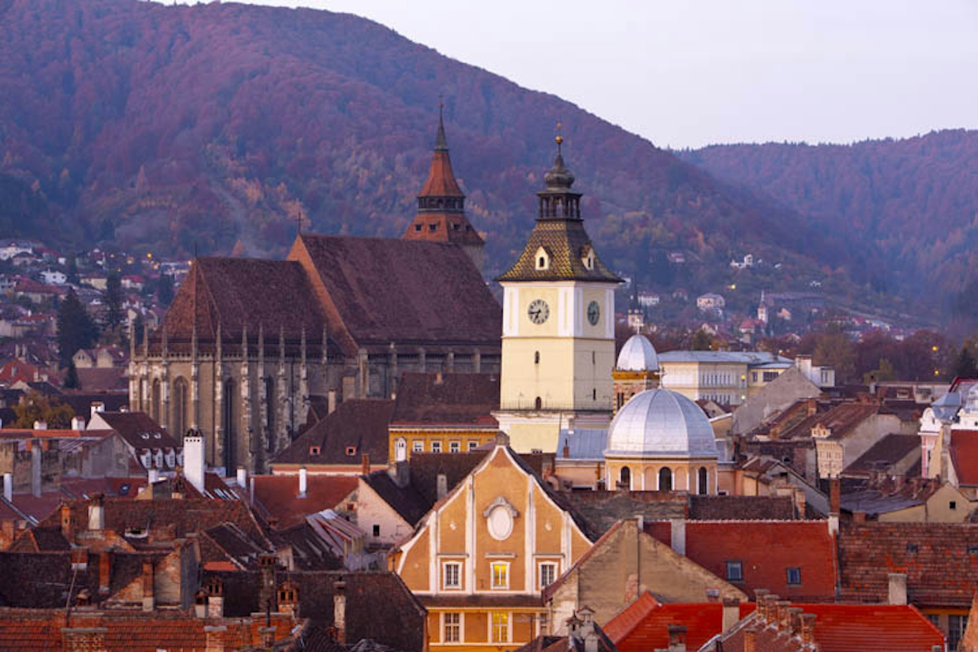 The gothic outline of Brasov's Black Church and silver-domed buildings in Brasov, Romania, in front of the thickly forested Carpathian Mountains.