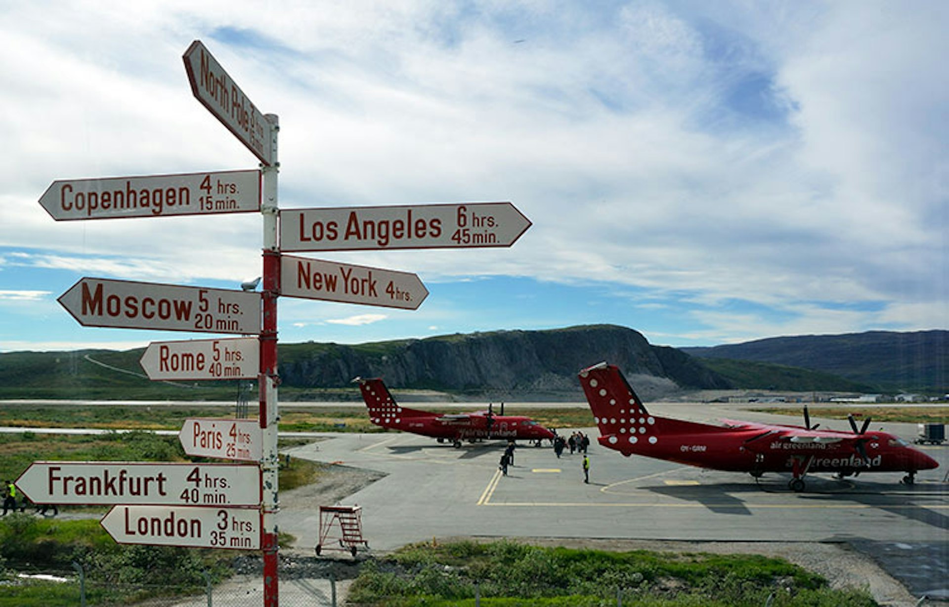 A sign pointing to a number of international destinations, including Cophenhagen, LA and London, with a background of red Air Greenland planes, against a rocky backdrop in Kangerlussuaq, Greenland
