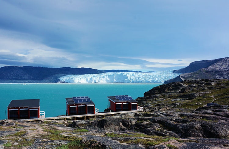 Three red huts with solar panels on the roofs, overlooking a bright aqua-coloured body of water with a white glacier in the background, under a cloudy sky.