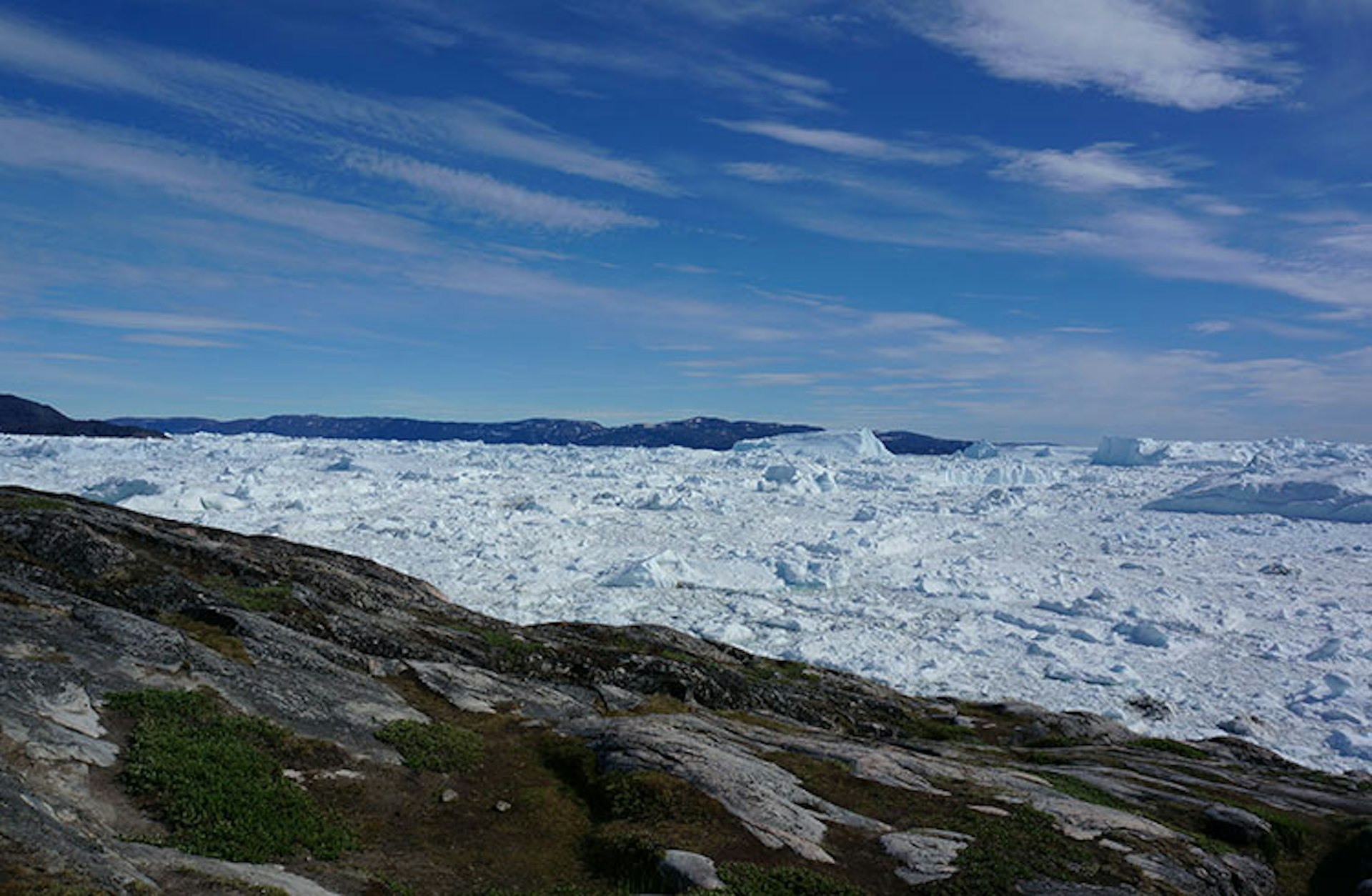 A vast expanse of ice, the Jakobshavn Glacier, stretches out under a blue sky, with a rocky, moss-covered hiking trail in the foreground