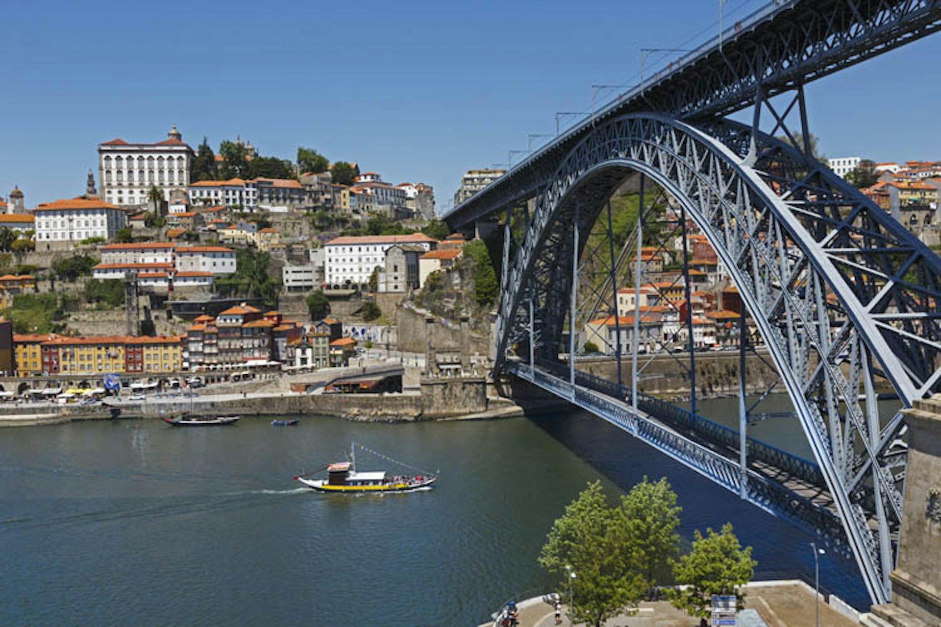 A small yellow boat glides under the metallic Dom Luis I bridge over the Douro River in Porto, Portugal; the far bank has a cluster of white and yellow blocks of flats.