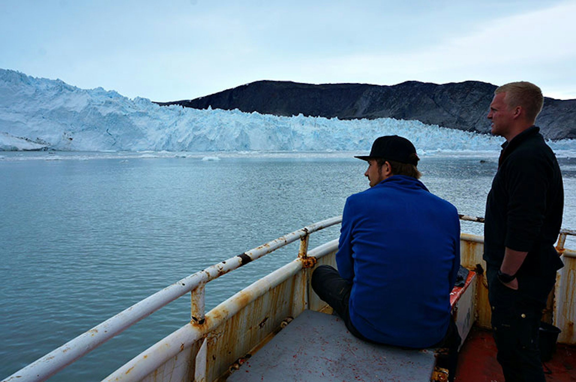 Two tourists stare out from the deck of a boat towards the large white face of a glacier in Greenland