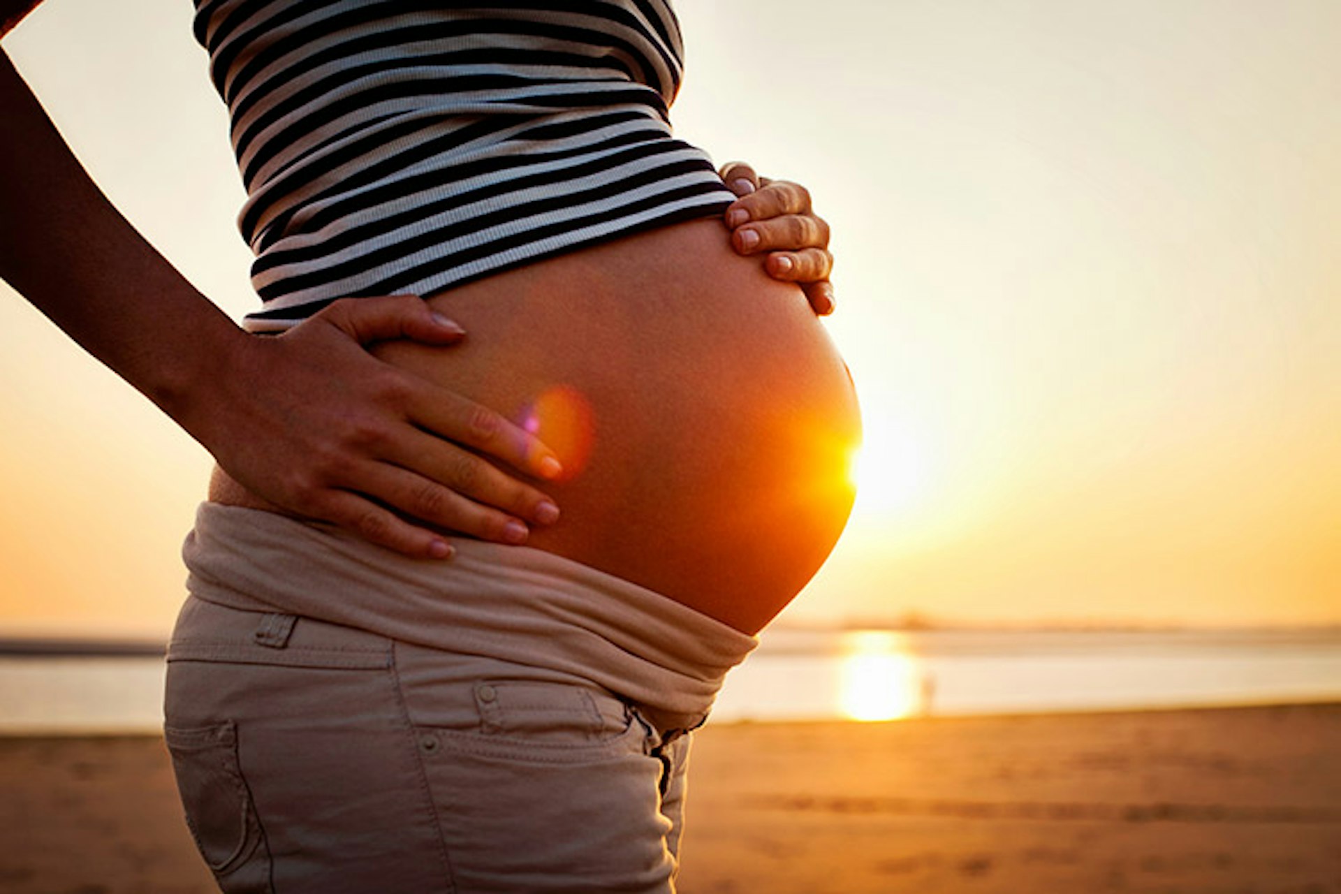 An image of a woman on the beach showing her baby bump as the sun sets in the background