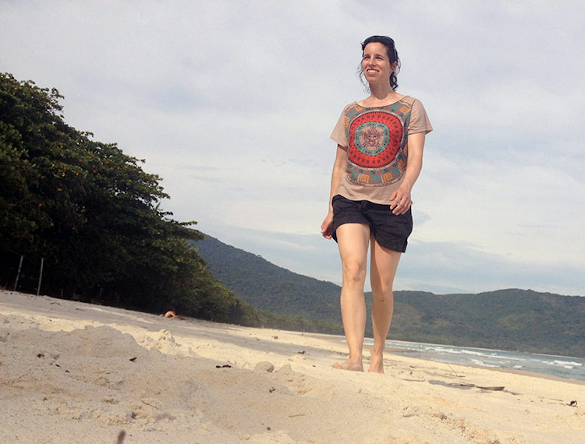 A woman wearing shorts and a t-shirt walks along a sandy beach approaching the camera