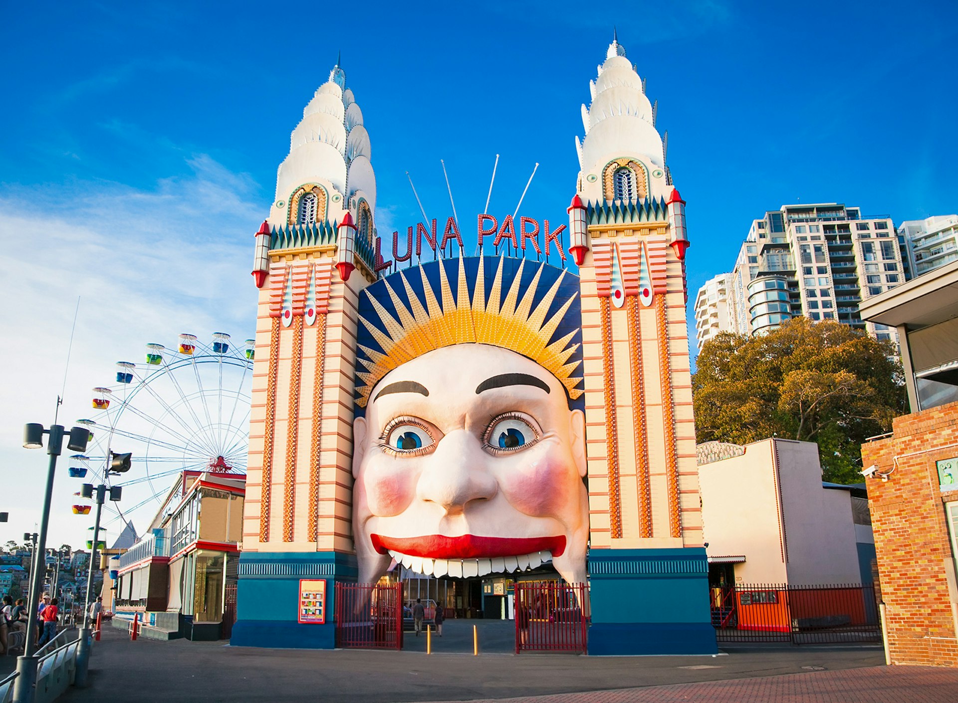 The Luna Park entrance has a large face smiling on the front, and people walk into its mouth to enter. Sydney, Australia