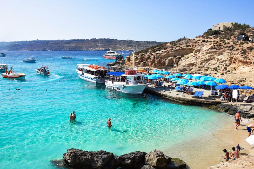 Η Blue Lagoon της Μάλτας, μια παραλία με ρηχά τιρκουάζ νερά, στην οποία λίγοι άνθρωποι κολυμπούν ενώ άλλοι κάθονται στην άμμο. Πιο έξω μερικά σκάφη είναι αγκυροβολημένα.