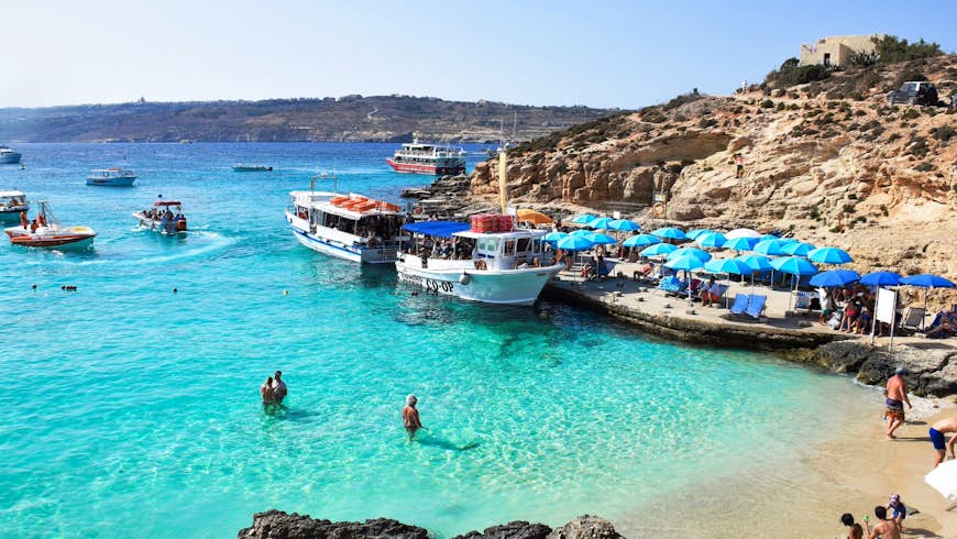 Malta's Blue Lagoon, a beach with shallow turquoise water, in which a few people swim while others sit on the sand. Further out a few boats are moored. 