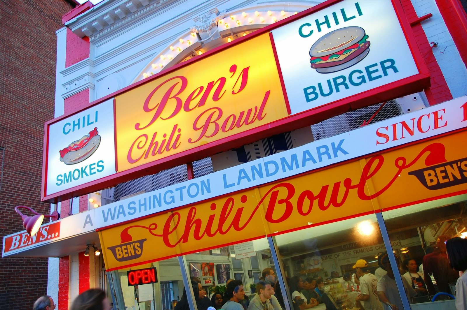 exterior shot of Ben's Chili Bowl, a diner in DC with colorful red and yellow vintage signage. Behind the glass window, the restaurant is crowded with patrons of all ages; weekend in washington, DC