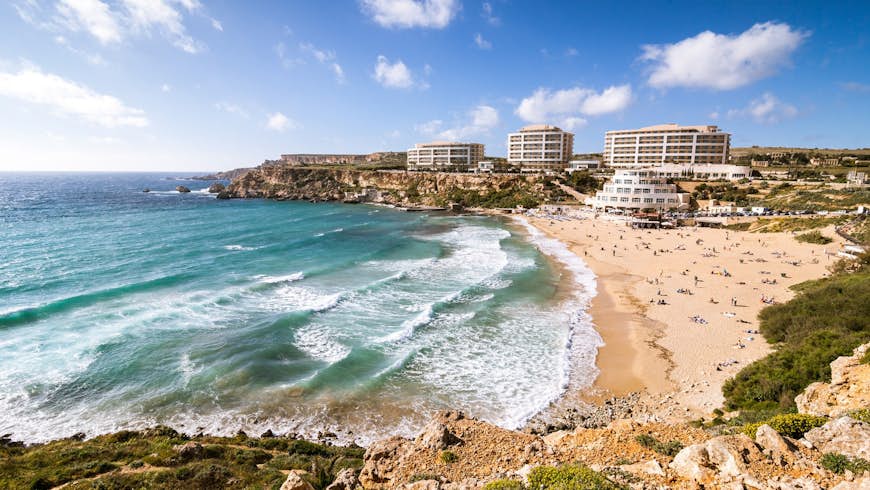 A sweeping view of Golden Bay in Malta with large waves lapping the beach, which is packed with sunbathers. In the distance a few hotels are perched on the top of the hill
