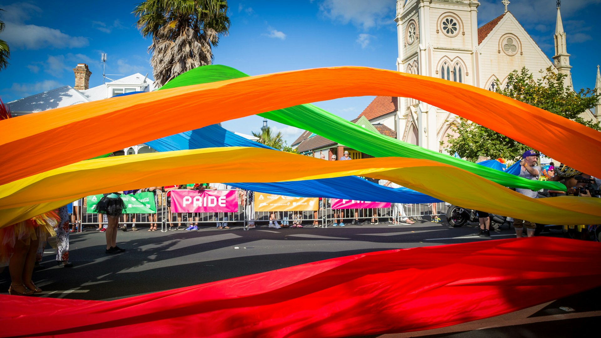 Banners of material in every colour of the rainbow are being waved across a street; in the background are a crowd of onlookers standing in front of a church.