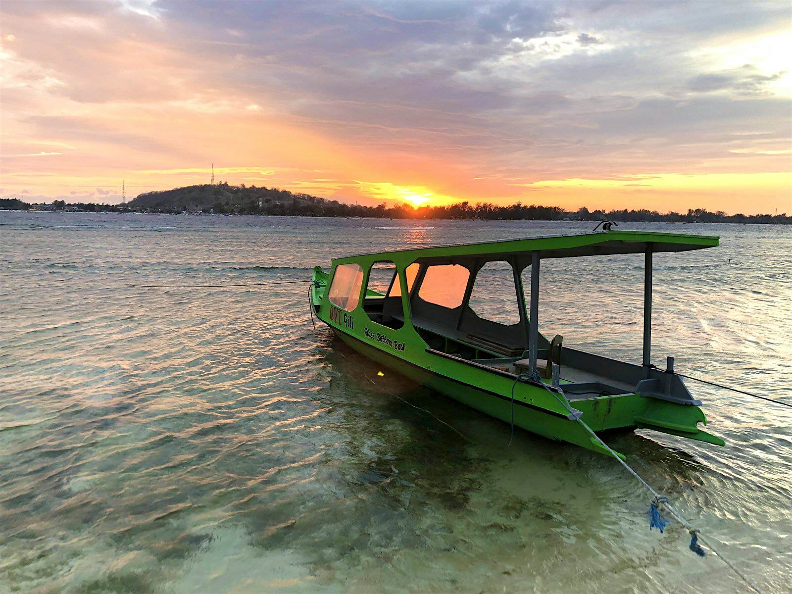 Gili Islands Gili Meno A small green boat is moored in shallow blue water with a colourful sunset and another island in the distance