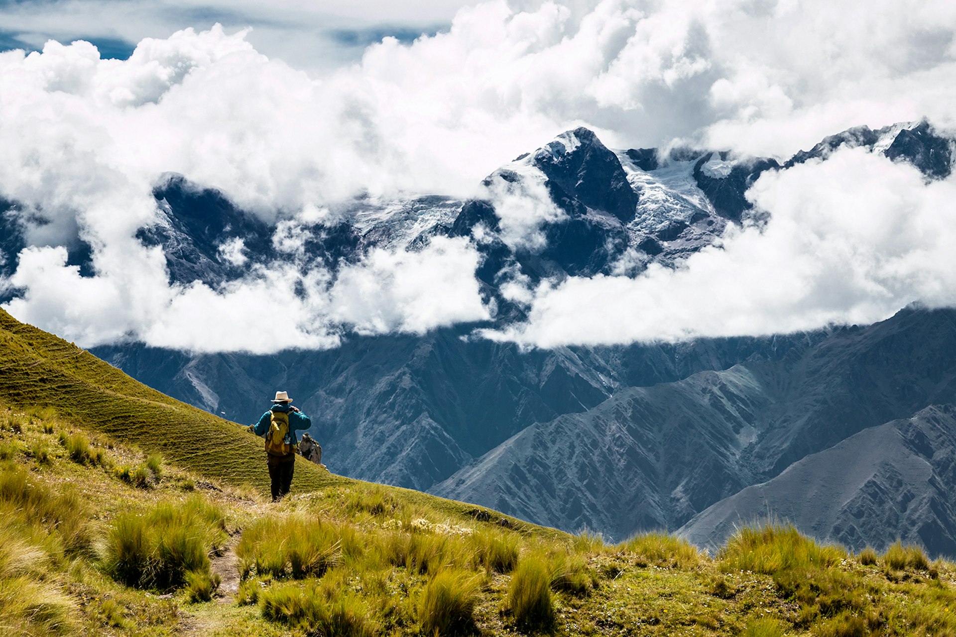 A hiker walks on a green hillside with grey mountains and clouds in the background © Pintai Suchachaisri / Getty Images