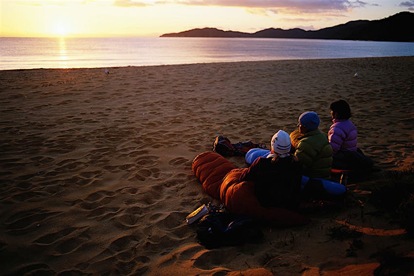 Three people sit on a sandy beach in sleeping bags watching the sunset