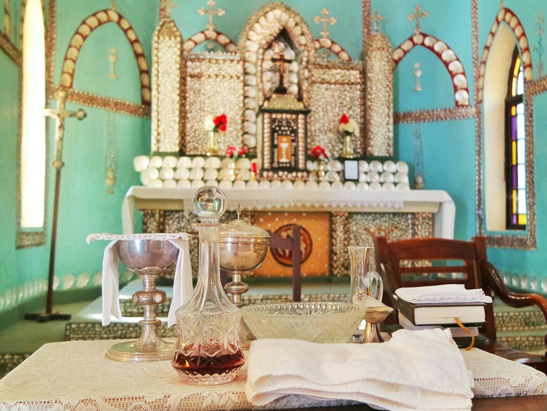 Church interior with chalice, bible and wine decanter in the foreground, altar decorated with shells in the background and bright teal walls.