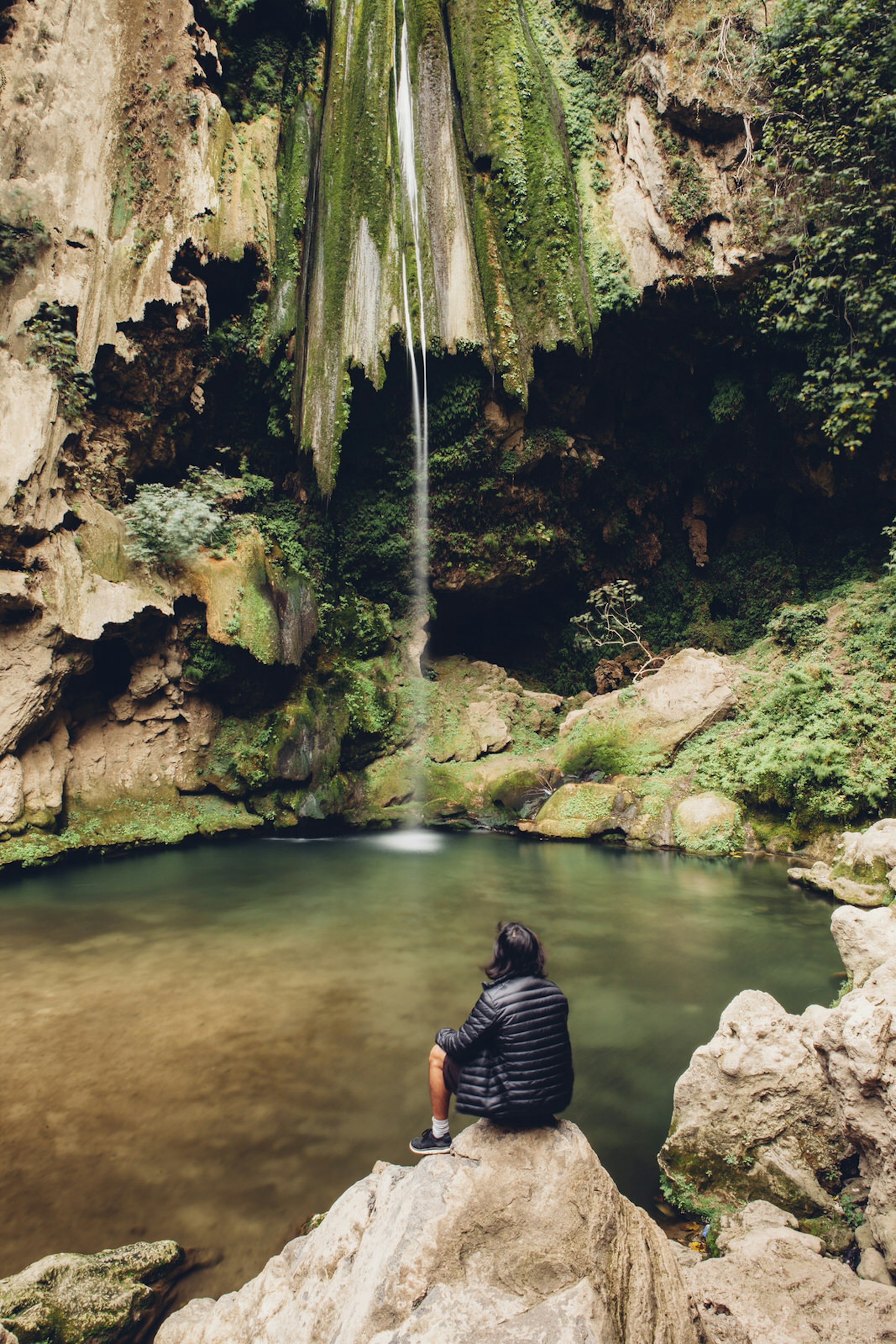 Features - High angle view of male hiker looking at waterfall while sitting on rock formations
