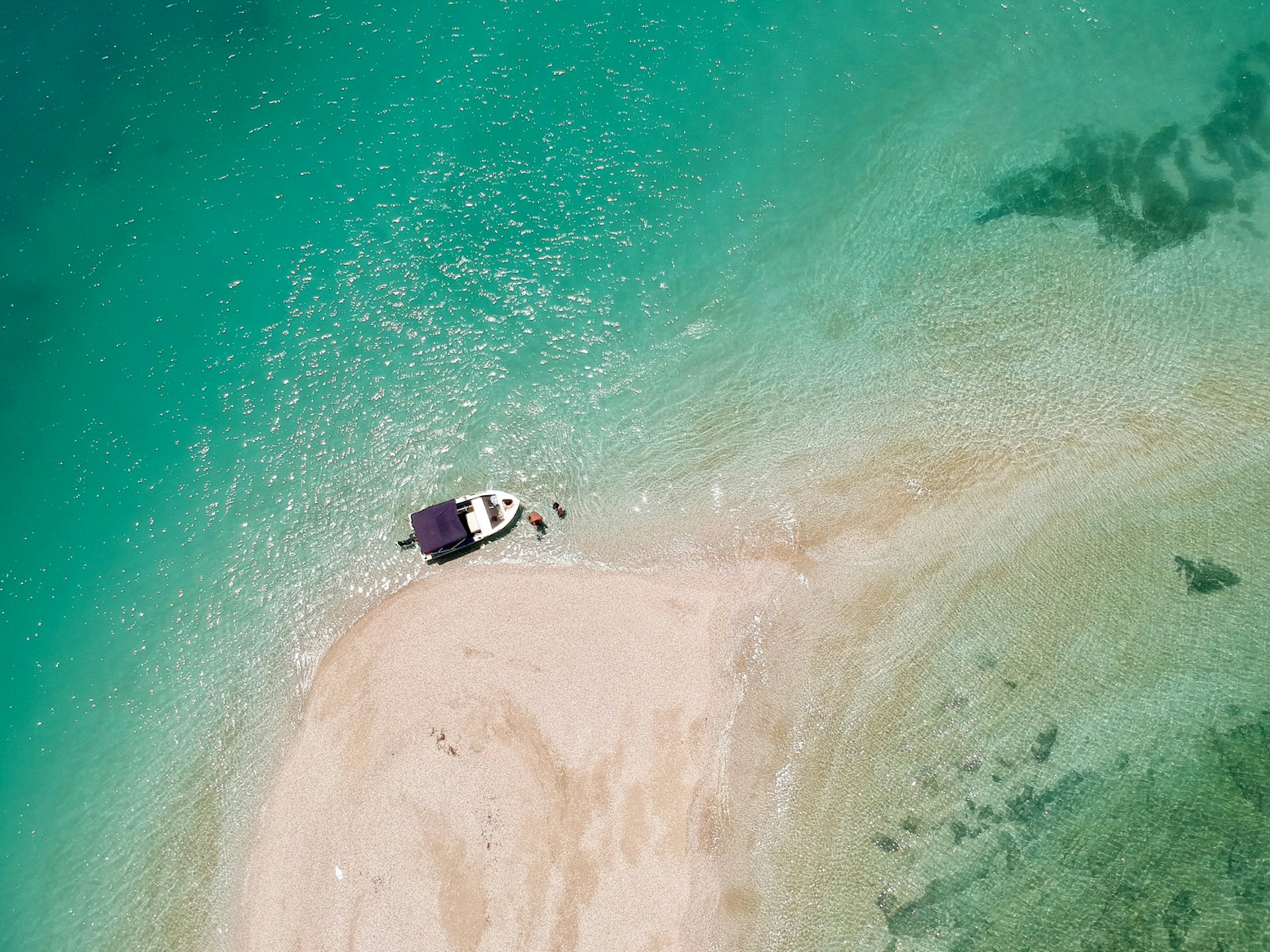 An aerial view of a boat moored on a spit of white sand and turquoise water with a couple of people standing in the water