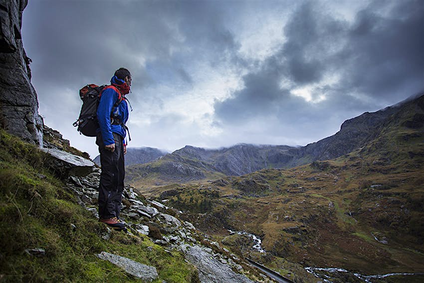 A man in hiking gear carrying a backpack stands on a rocky hillside surveying grey clouds that are rolling down the valley