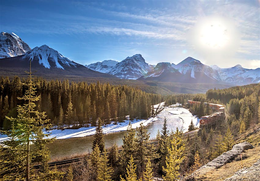 A red freight train curls around Morants Curve in Banff National Park; the track is surrounded by pine trees and there are snow-capped mountains in the background