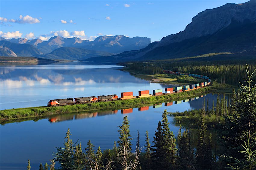 A freight train cruises through Swan Landing near Brule, Alberta; the train is reflected in the still waters of the lake while mountains are visible in the background