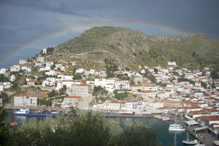 Many white buildings with red roofs line the bottom of a hill. There's a small harbour in the foreground and a rainbow over the hill