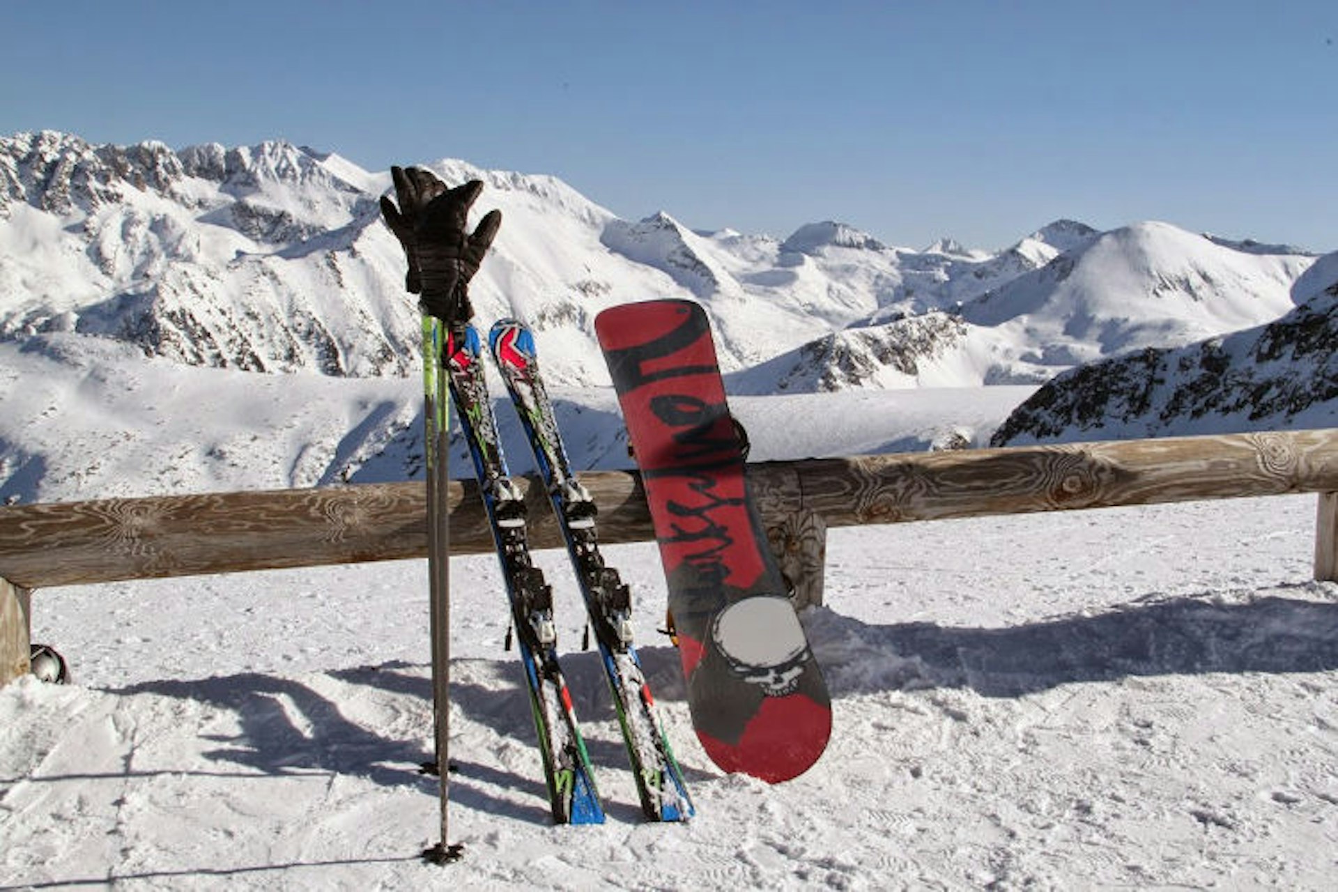 A set of skis and a snowboard lean against a fence in a mountainous snowy area