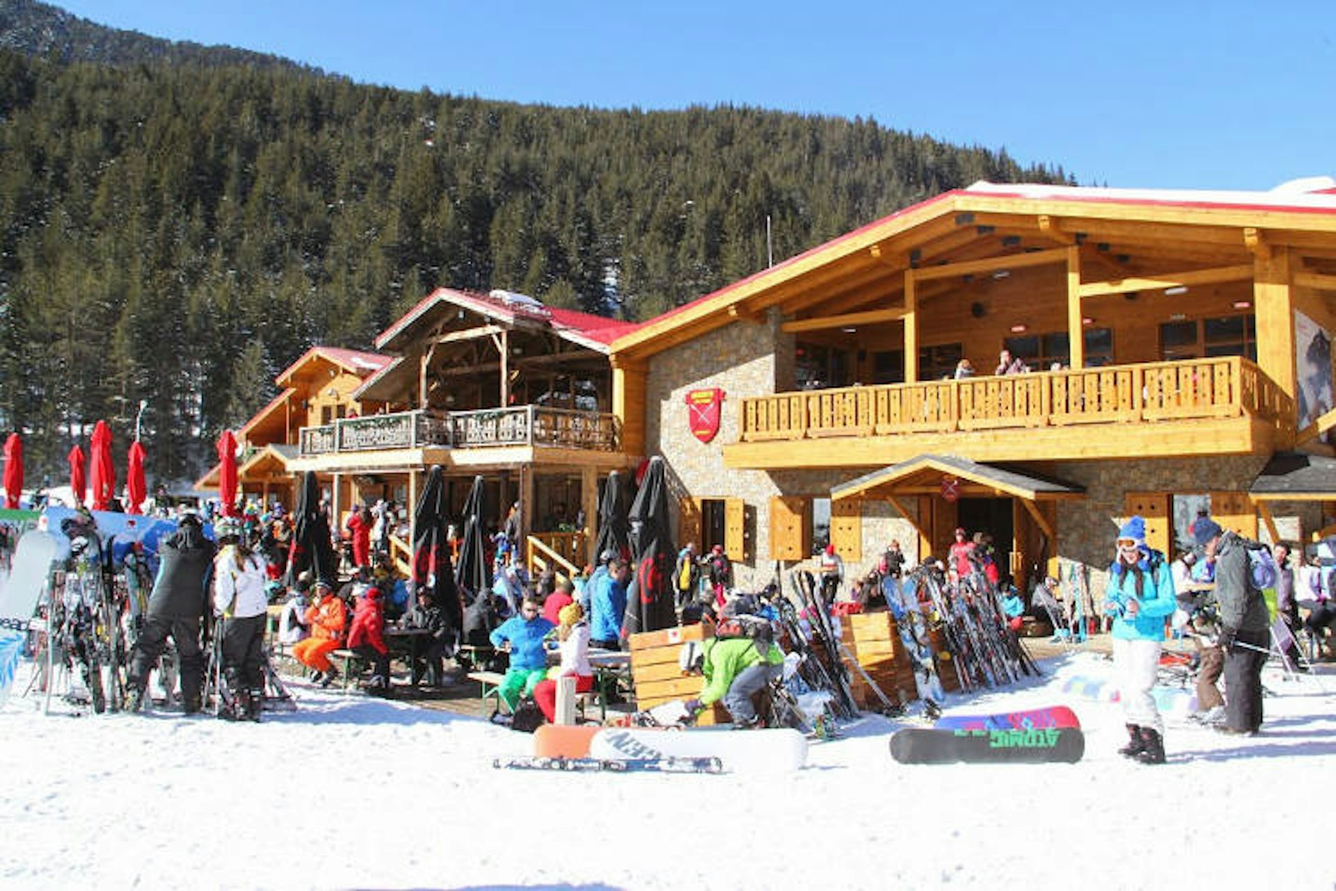 Three wooden chalet-style buildings with many people wearing colourful ski gear gathered in front