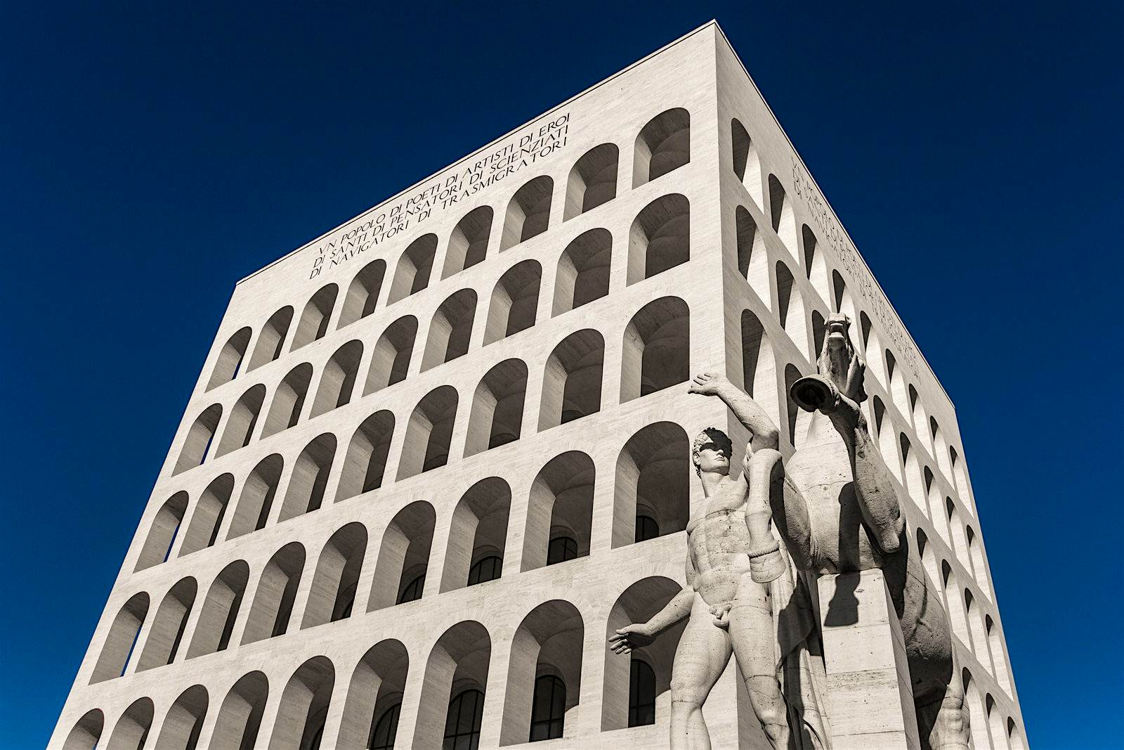 Palazzo della Civiltà del Lavoro, Rome A naked Roman marble statue, with his arm raised in a way that looks like he is shielding his eyes from this day's sun, stands in front of a modern cubic building with dozens of arches; 'The Square Colloseum'