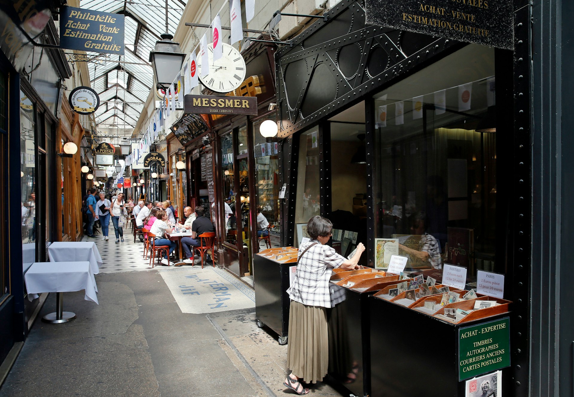 A lady looks through some wares of the Passage des Panoramas, while others stroll along or sit in chairs outside cafes.