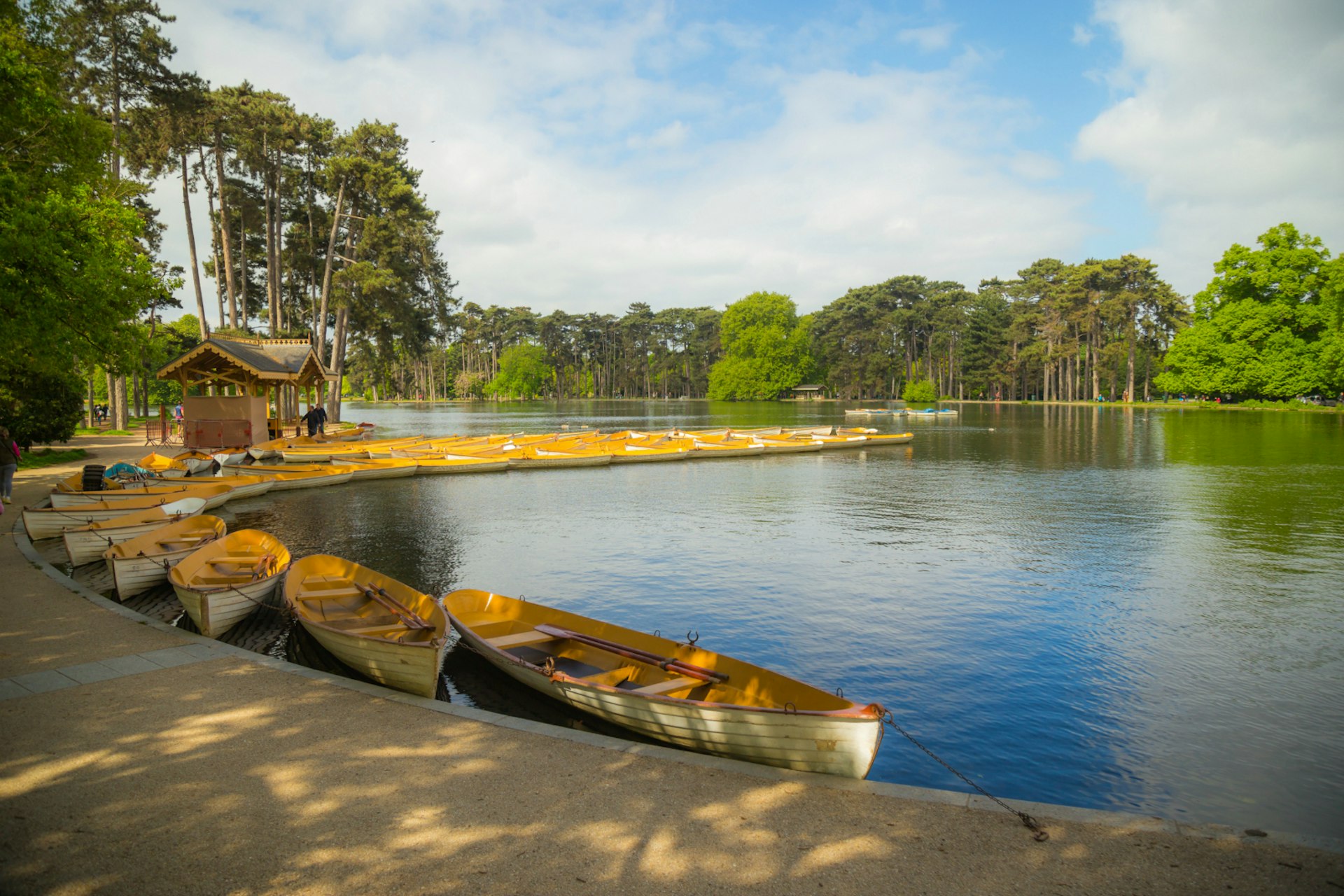 A number of yellow and white boats are moored on the banks of Lac Inférieur in Bois de Boulogne, Paris. The lake is large and surrounded by greenery.