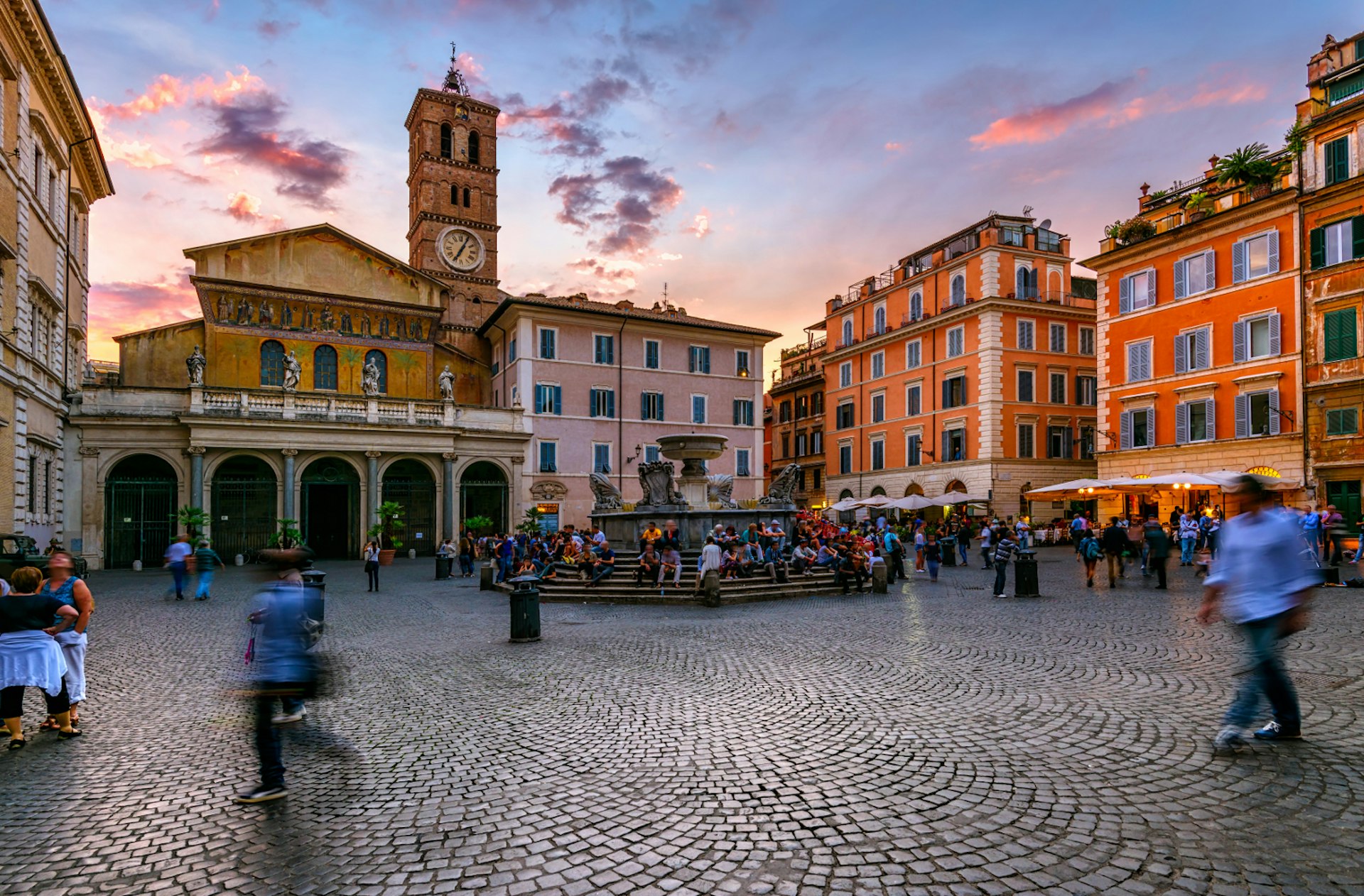 A cobbled square lined by historic buildlings, including Basilica di Santa Maria with its clock tower. People walk to and fro, while others sit on the fountain.
