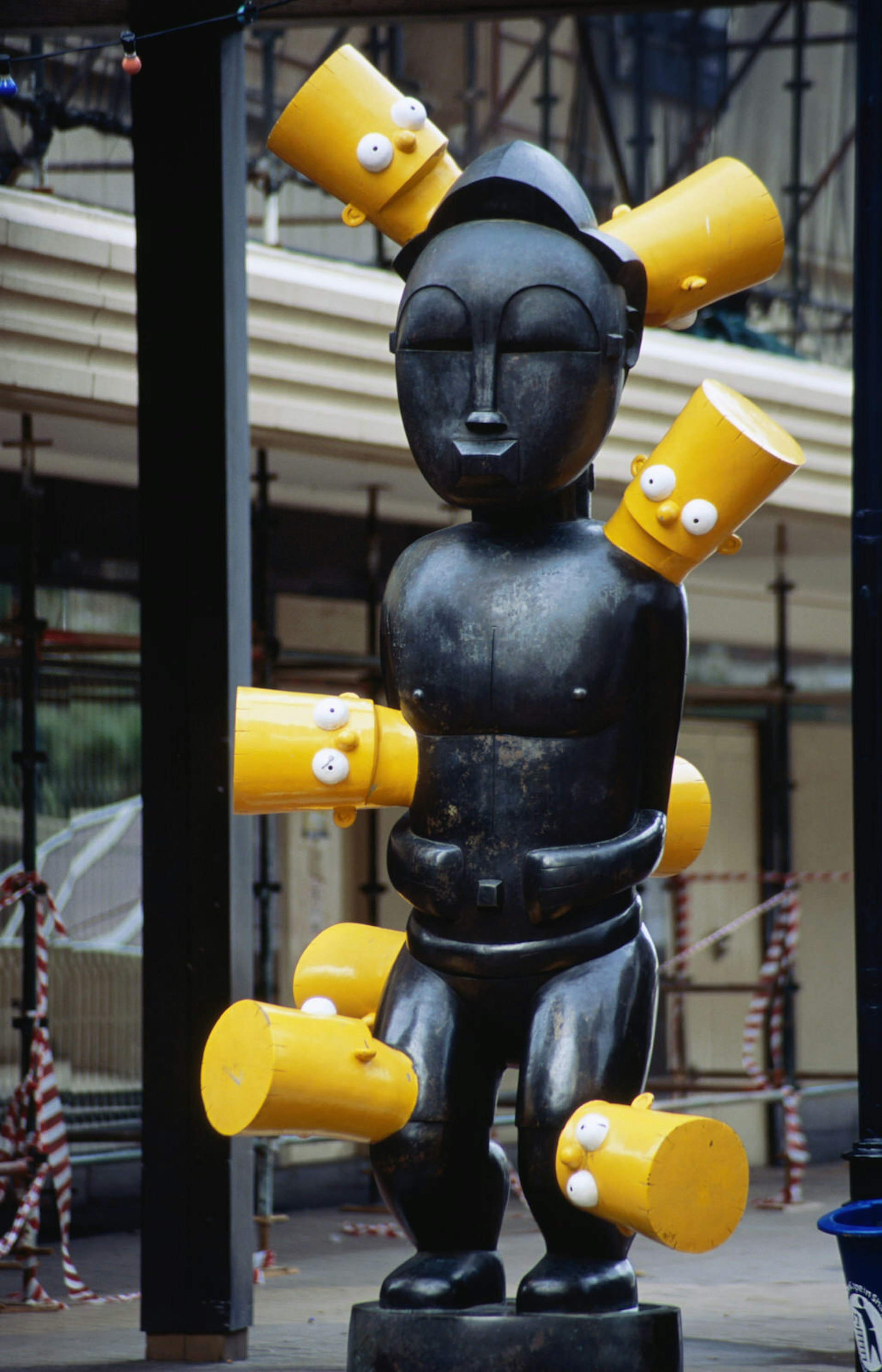 A black bronze sculpture of a human that resembles a traditional West African carving is dotted with bright yellow Bart Simpson heads