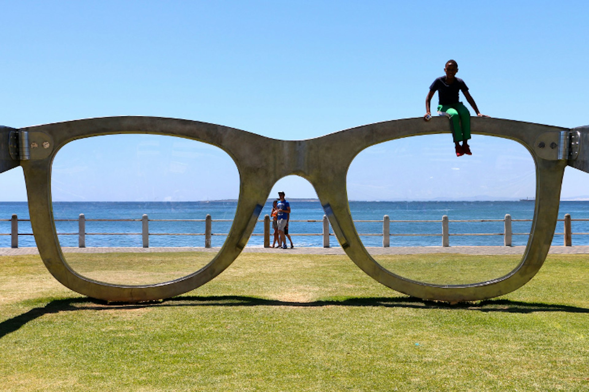 A huge pair of Ray Bans sit on the grass, with the sea in the background; a young person sits atop one of the framesPerceiving Freedom