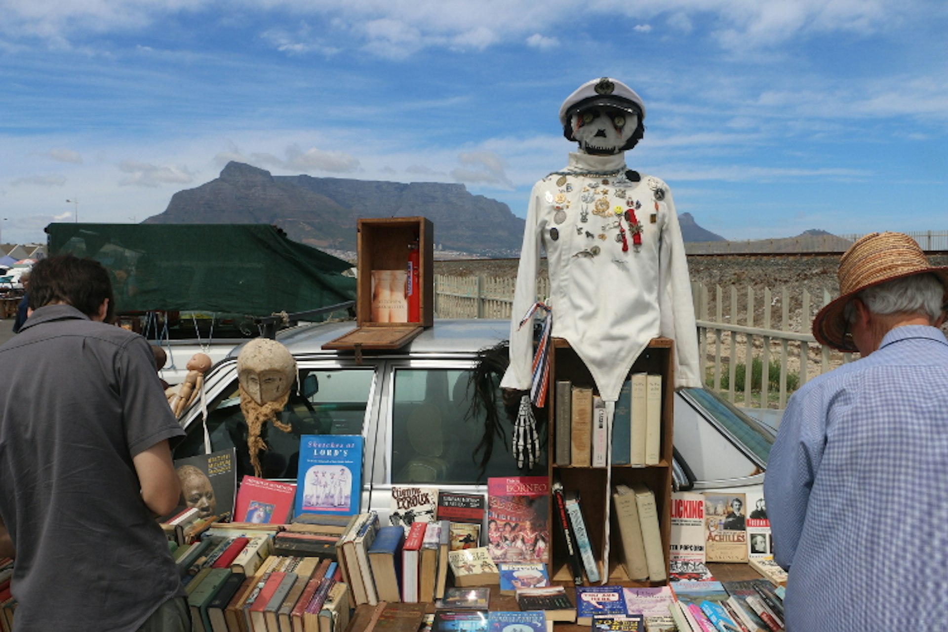 Shelves of books and a make-shift scare-crow wearing a skippers hat and white shirt covered in decorative pins stand in front of a car at the market; Table Mountain is the background.