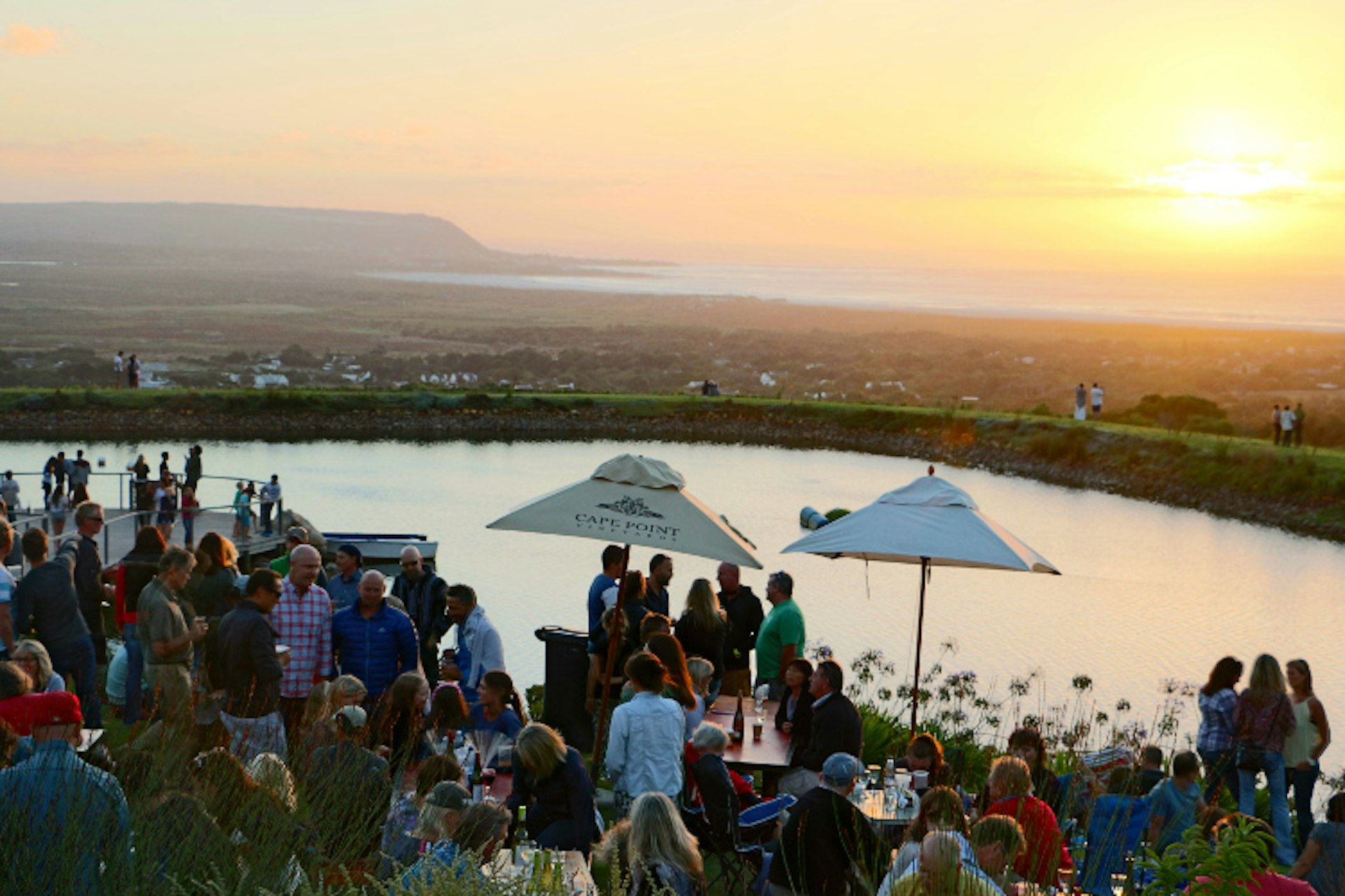 A crowd of people stand on a deck with tables and several large sun umbrellas at sunset; a water reservoir sits just behind, with the ocean in the distance.