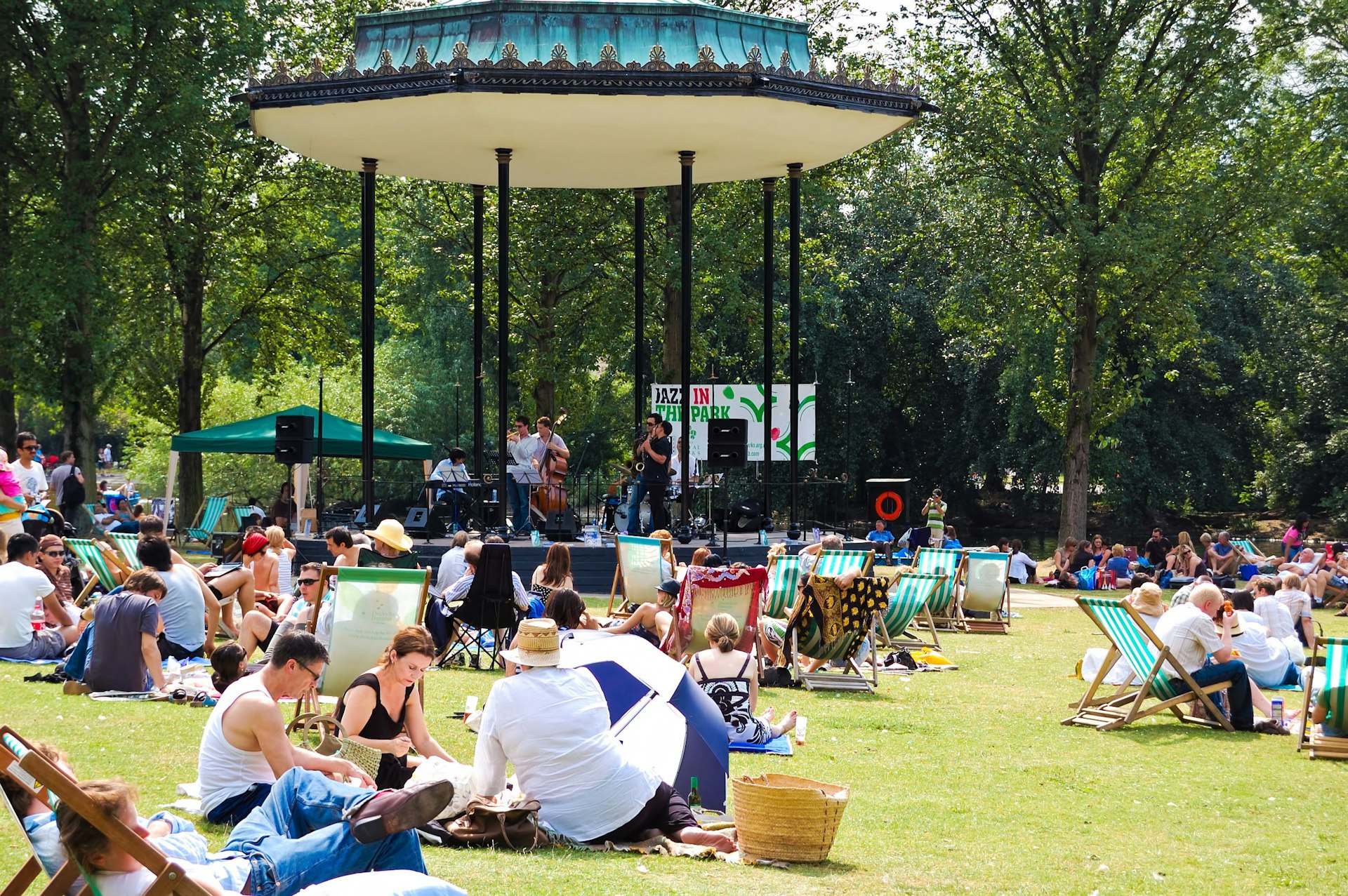 People sitting in a London park in the sunshine, while a jazz band performs in a bandstand.